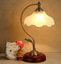 Vintage-style table lamp with a warm glow, next to a small clock and figurine on a beige surface.